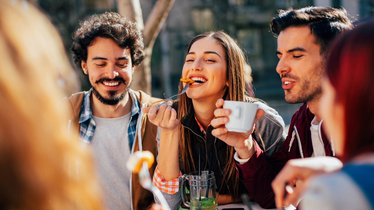 Friends around an outdoor table in the sun, sharing food and coffee.