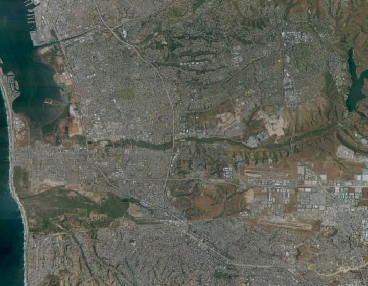 An aerial photograph of Chula Vista and the South Bay. San Diego Bay and the marina run down the western side, two freeways run north to south through the city, master-planned housing and open land lie to the east around the Otay reservoir, and the border with Mexico crosses the bottom of the frame.