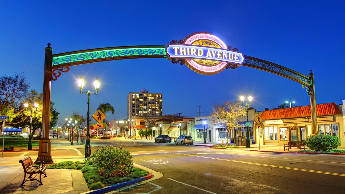 The lit Third Avenue arch over the street at dusk, with shopfronts and streetlights beneath it.