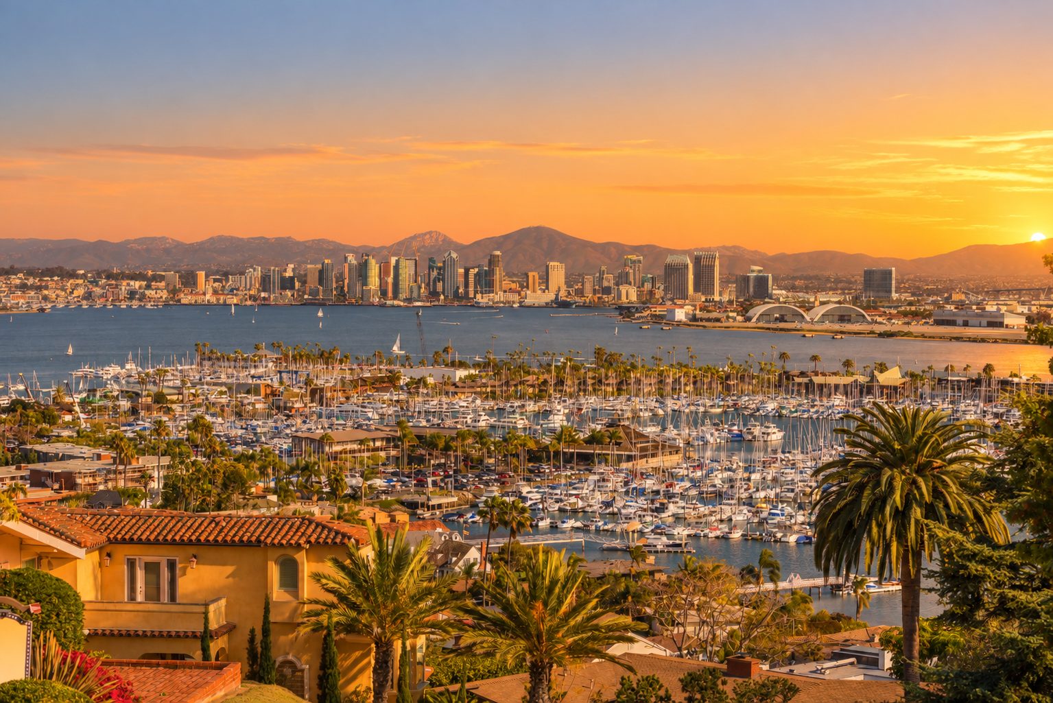 A marina packed with moored yachts at sunset, a city skyline along the far shore and hills on the horizon behind it.